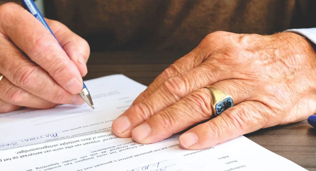 Close-up of a senior adult signing a legal document with a focus on hand and gold ring.