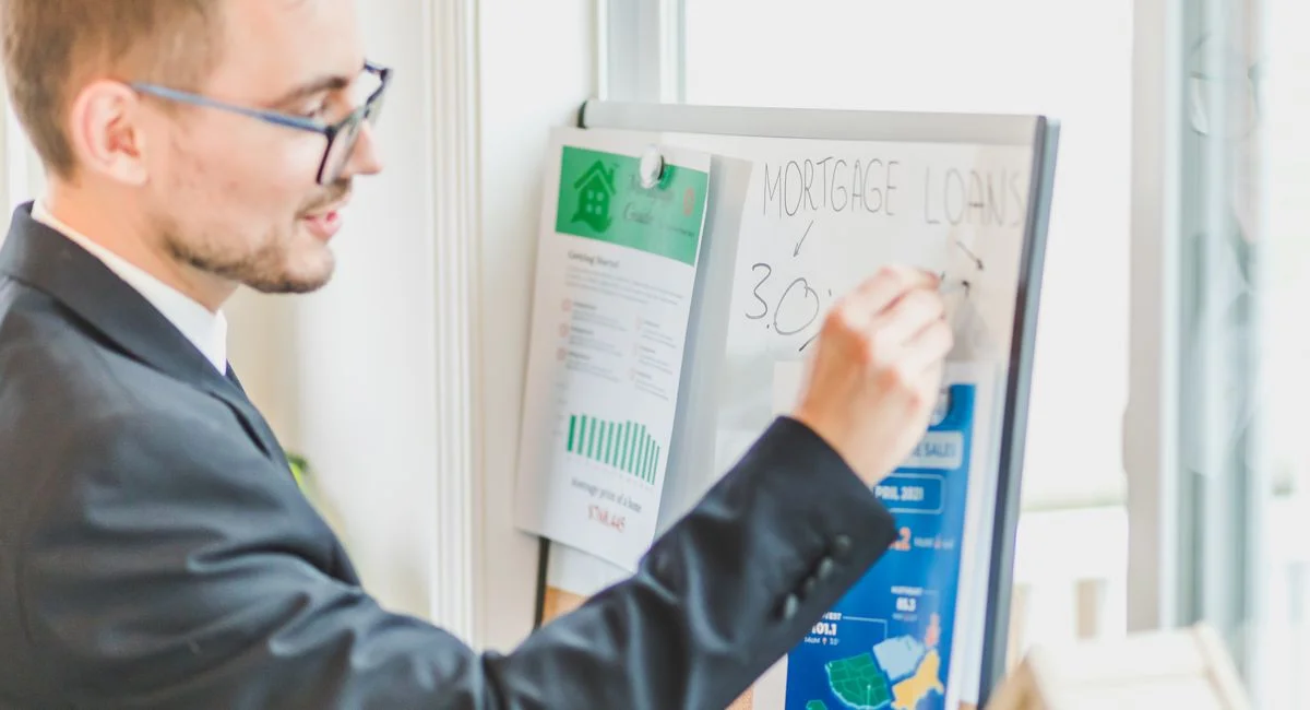 Businessman in suit writes on whiteboard displaying mortgage loan rates during a real estate meeting.