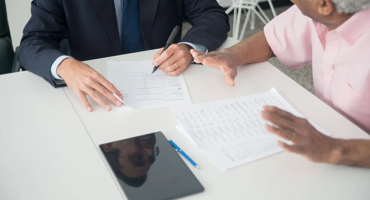 A business meeting between an elderly client and a consultant discussing documents at an office table.