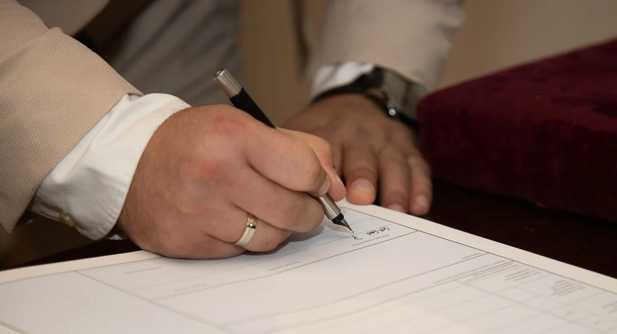 A close-up of a man signing a document, showcasing a wedding ring and pen.