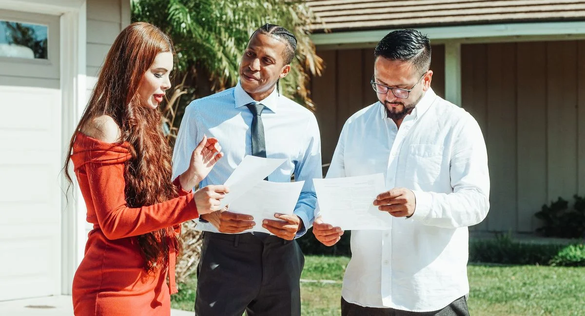 A diverse group evaluates real estate documents outside a residential property in daylight.