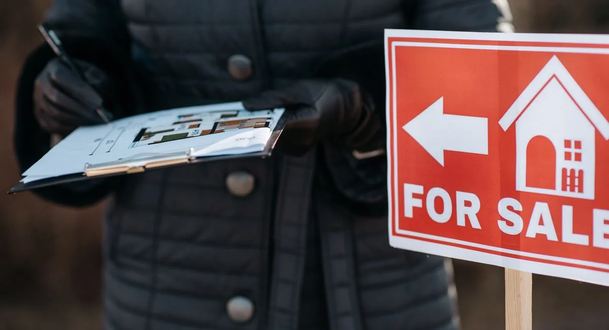 A real estate agent reviews plans next to a 'For Sale' sign outdoors, indicating property availability.