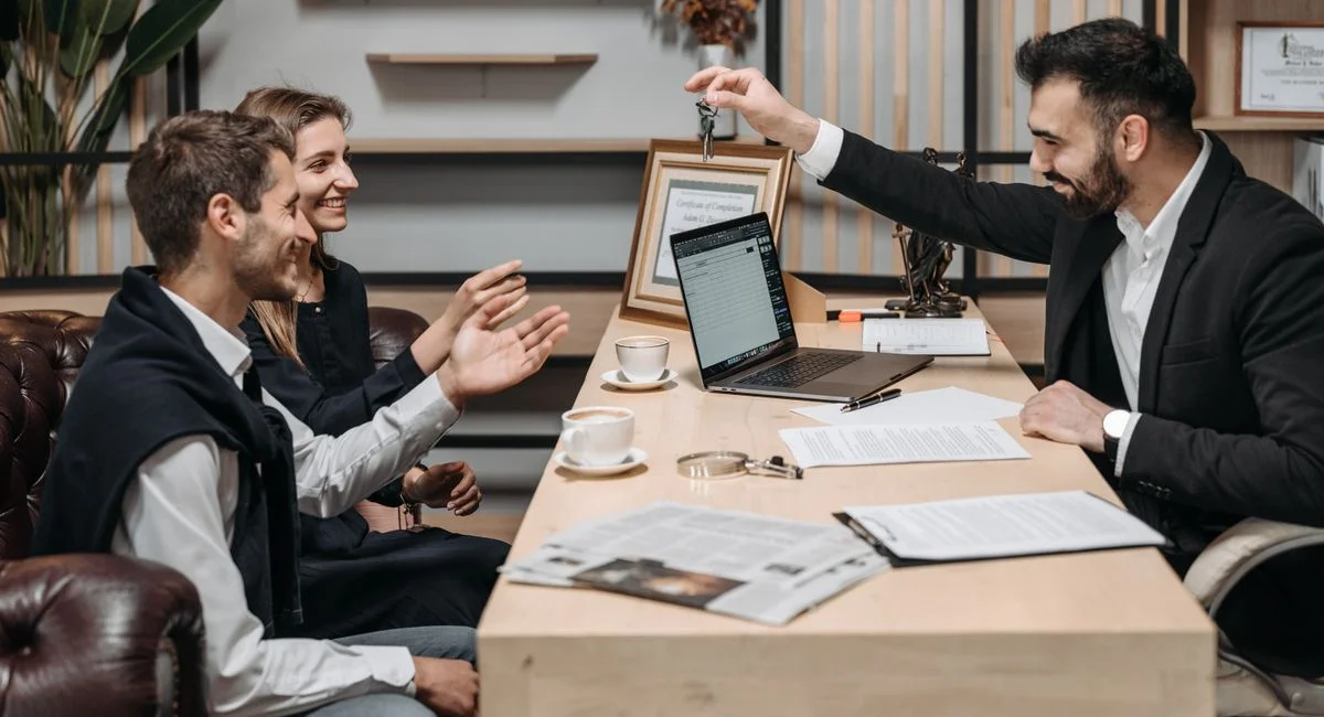 Business professional handing keys to smiling clients at an office desk during a consultation.