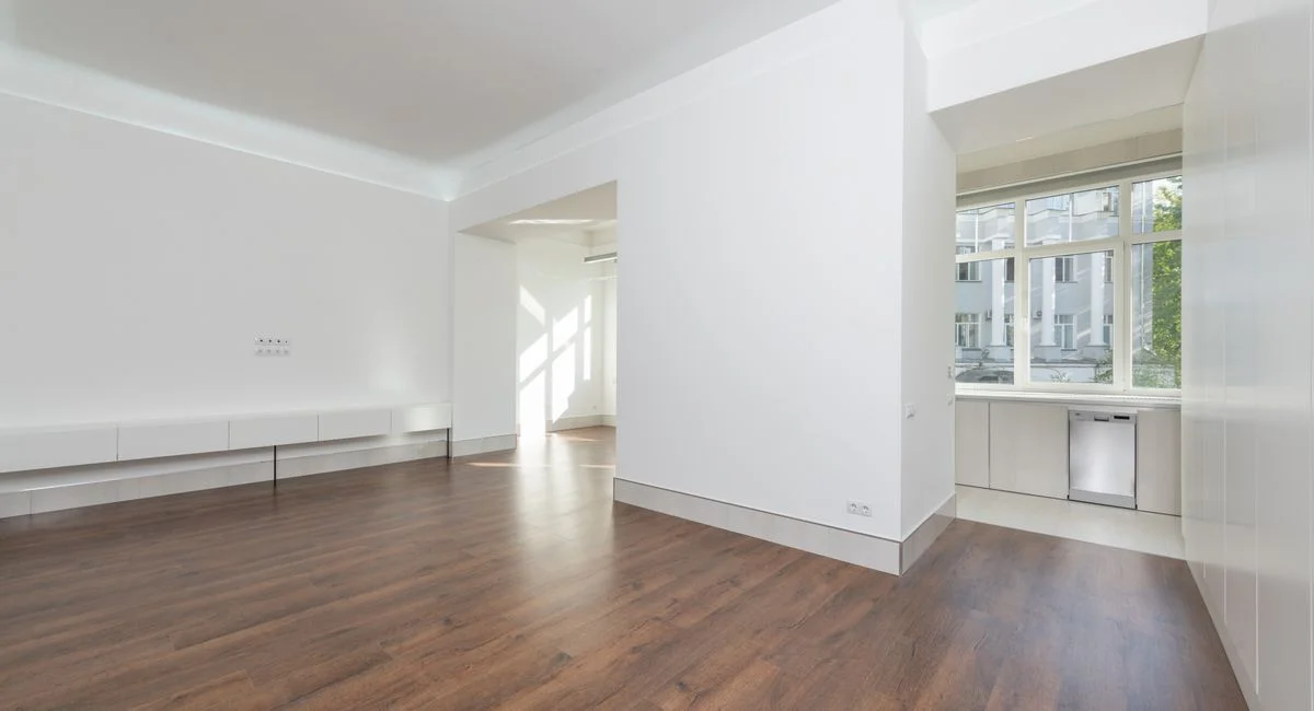 A contemporary empty apartment with white walls and wooden flooring, featuring large windows.