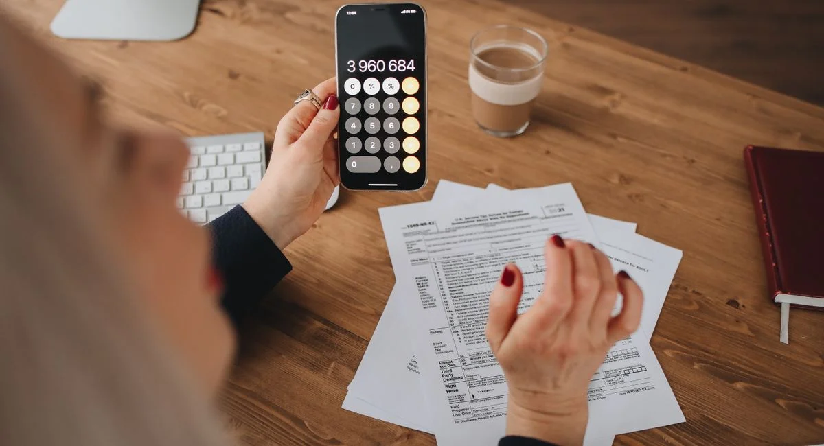 A woman calculates on her smartphone while working with documents at a desk.