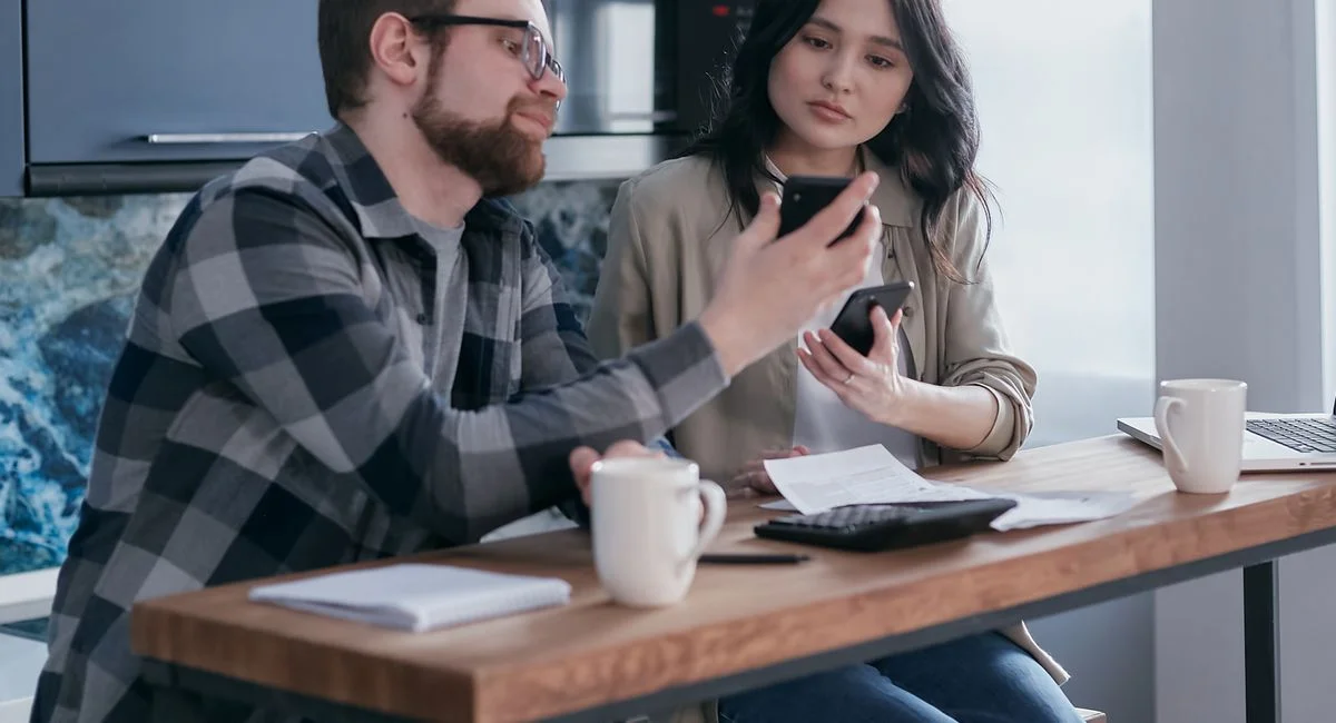 Young couple discussing finances using mobile phones at a kitchen table.
