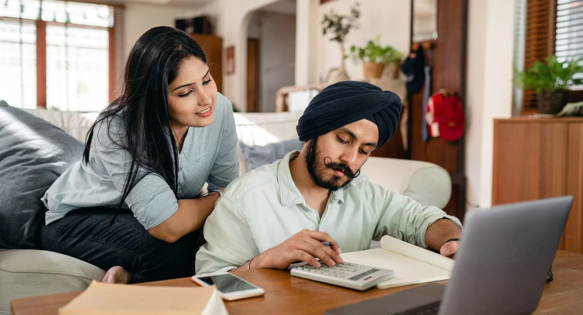 Smiling focused ethnic woman sitting on sofa at home watching working ethnic husband calculating sitting at coffee table amon