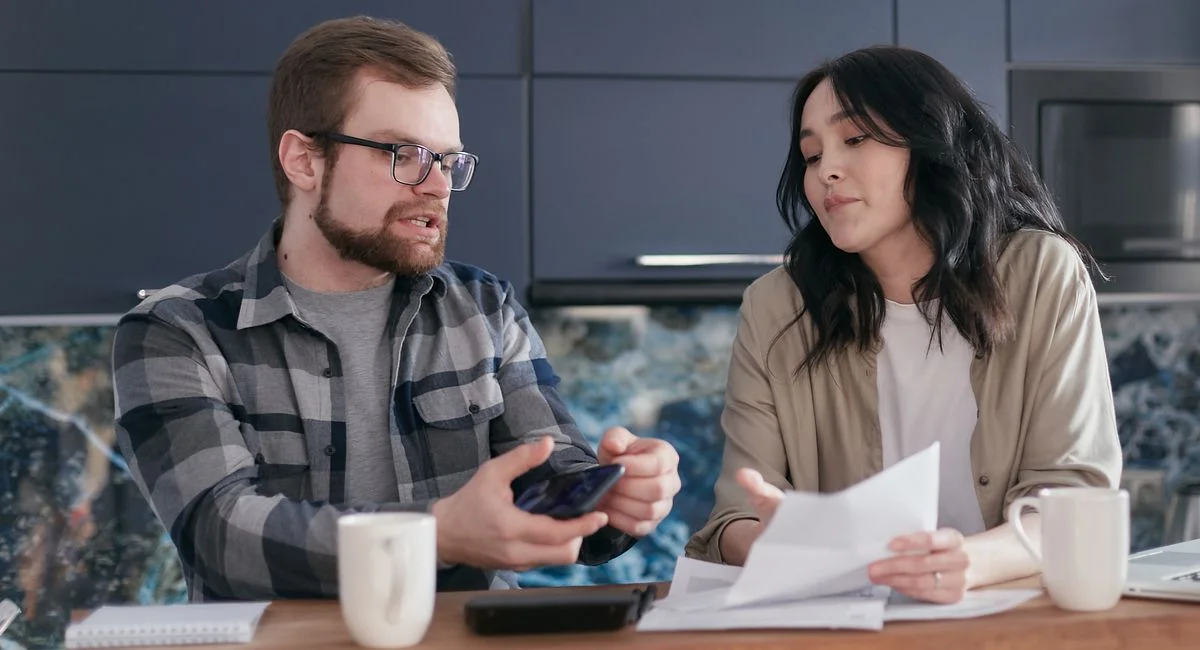 A couple sits at a kitchen table reviewing documents and discussing finances.