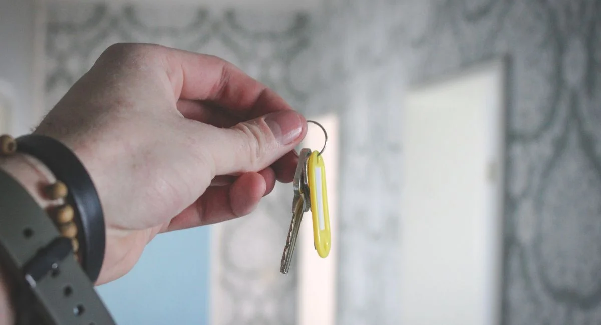 A hand holding a key against a blurred background of patterned wallpaper indoors.
